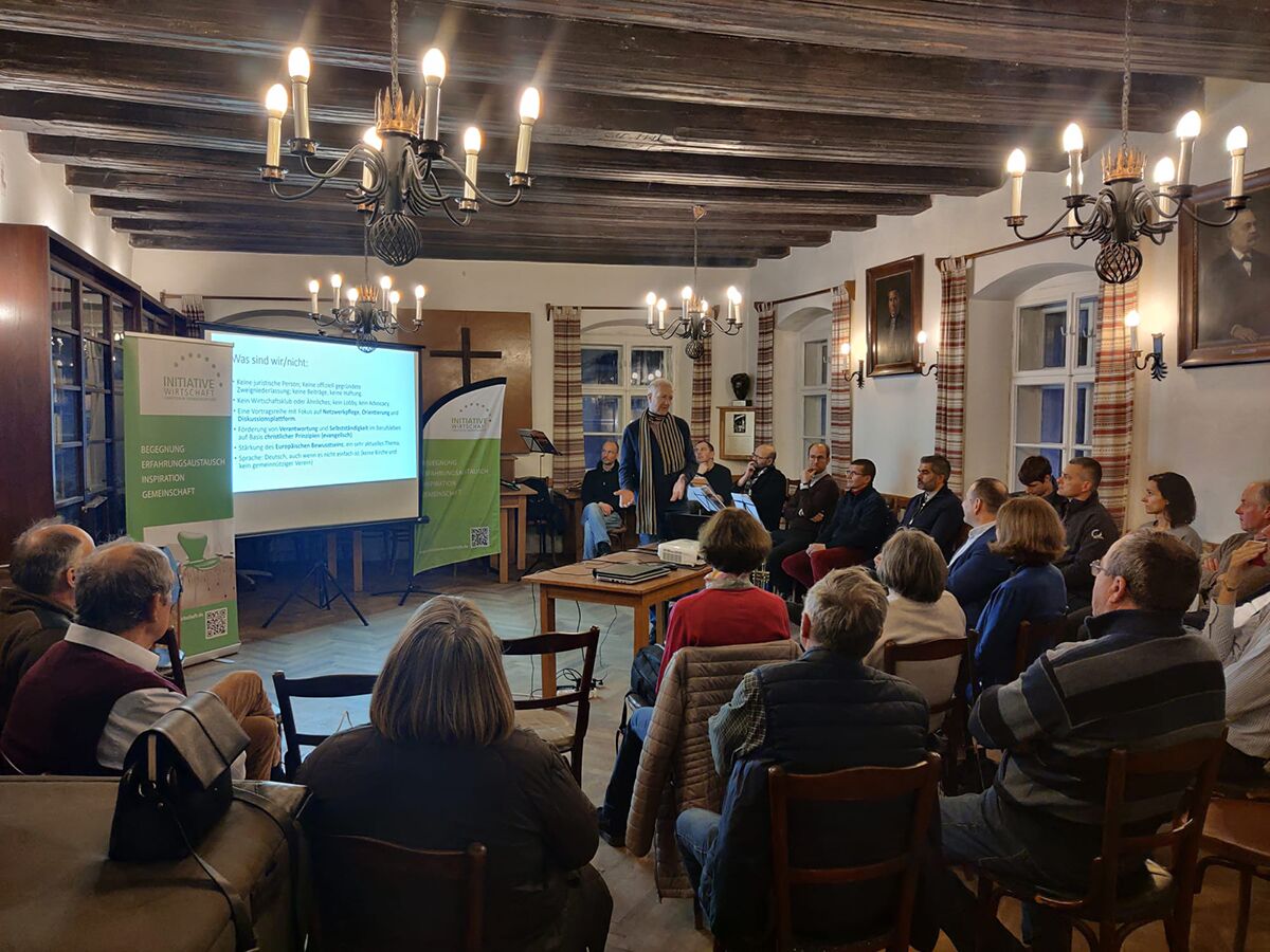 Gut zwanzig Teilnehmer in einem Saal mit dunkler Holzbalkendecke und Kerzenleuchtern; vorne spricht ein Mann vor einer Leinwand, daneben zwei grüne Rollbanner der INITIATIVE Wirtschaft.